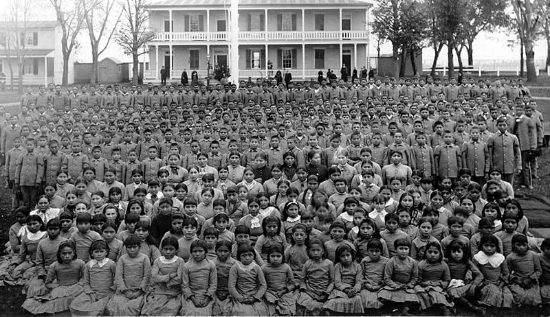 A group photo of hundreds of Native American children and teenagers, few if any of them smiling, lined up in front of a school building