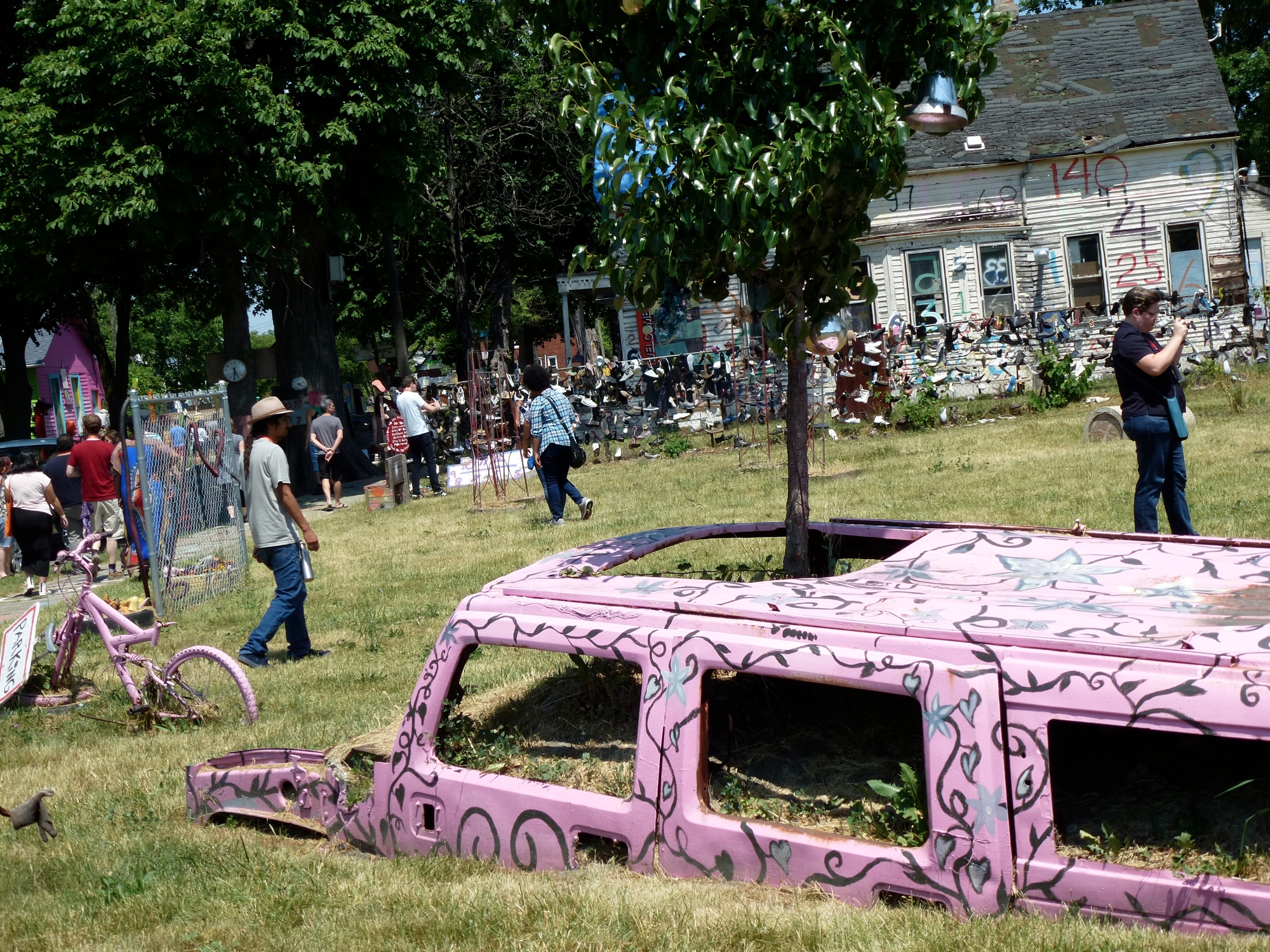 A Hummer appears to be half buried in the ground. Its windowless frame is painted matte pink with vine-like designs. A small tree grows out of its sunroof. In the background are a house, other outdoor art pieces, and people of various races and genders looking at them.