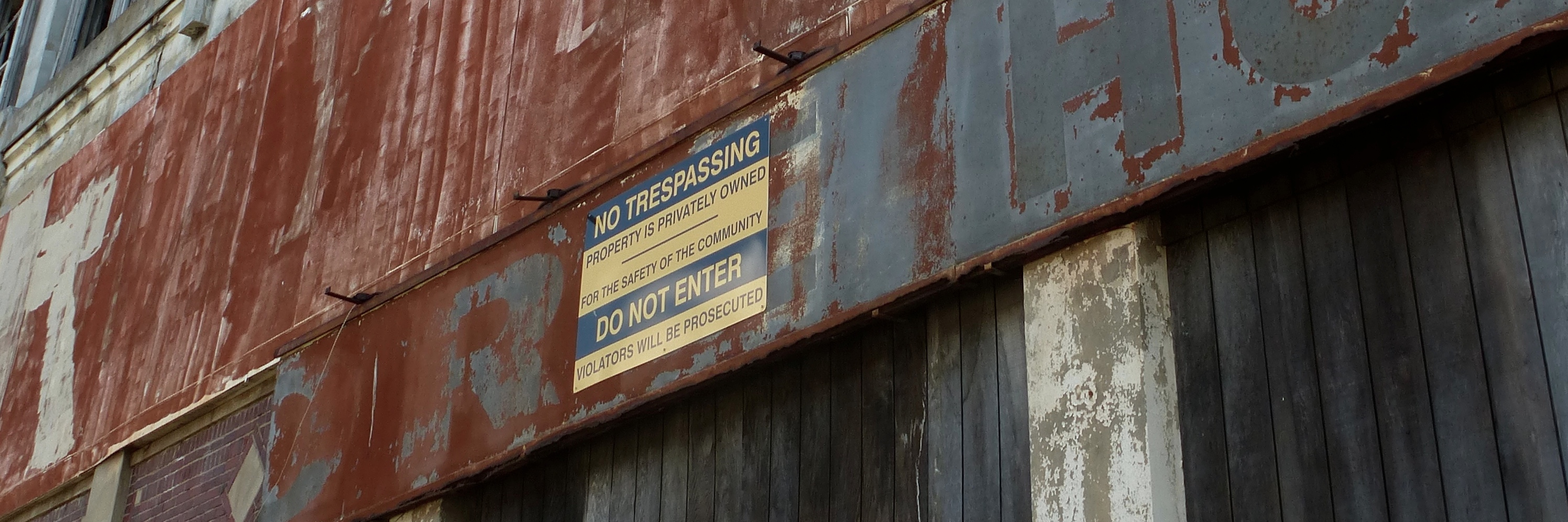 A section of the front of an abandoned building, with flaking paint, discolored wood, and rusty metal with large faded letters. Affixed to the metal is a yellow and black no-trespassing sign.